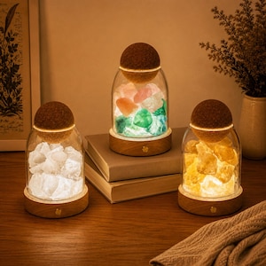 Può includere: Tre lampade a barattolo in vetro con basi in legno e coperchi in sughero. La lampada a sinistra contiene pietre bianche, quella centrale pietre multicolori e quella a destra pietre gialle. Ogni lampada emette una luce calda dalla base.