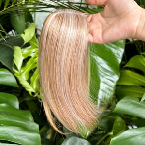 Op de afbeelding: Een haarstukje met steil, meerkleurig blond haar met lichte gouden en platina highlights. Het haar is glad en glanzend, met een natuurlijk ogende scheiding en een gelaagde snit die recht naar beneden valt in een uniforme, verzorgde stijl.