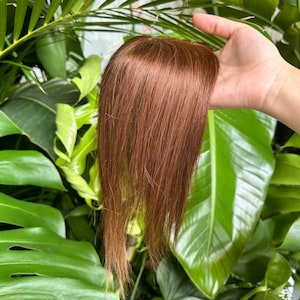 Puede incluir: Un postizo capilar con mechones lisos y suaves de color caoba. La pieza presenta una raya de aspecto natural y está diseñada para cubrir la zona de la coronilla, mostrando un tono marrón rojizo uniforme en toda su longitud.