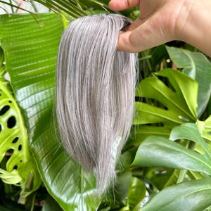 May include: A hair topper piece featuring straight, shoulder-length hair in a cool-toned silver and light gray color palette. The hair strands are smooth and sleek, showcasing a natural-looking texture with subtle variations in the gray tones.