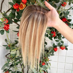 May include: A hair topper featuring straight, multi-tonal blonde and light brown highlights. The piece has a visible monofilament base at the crown, with smooth, layered strands that transition from a darker root color to lighter blonde ends.