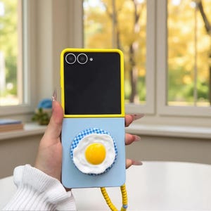 May include: A foldable smartphone case featuring a bright yellow frame and a light blue lower section. A circular grip attachment is centred on the bottom half, decorated with a blue and white gingham pattern and a 3D fried egg design.
