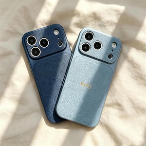May include: Two smartphone cases with a textured, leather-like finish. One case is dark navy blue, and the other is a lighter dusty blue with the name Ruby embossed in gold lettering. Both feature a raised camera module with three circular lenses.