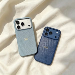 Puede incluir: Dos fundas de teléfono rectangulares con un patrón texturizado de piel de lagarto. Una funda es azul claro con las iniciales SR grabadas en oro, y la otra es azul oscuro con las iniciales MS grabadas en oro. Ambas tienen un recorte para la cámara.