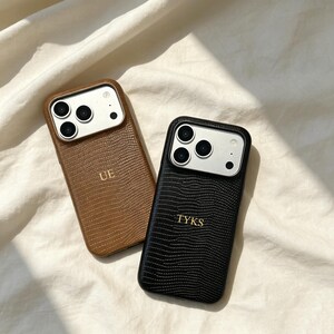 May include: Two smartphone cases with a textured lizard-skin pattern. One case is brown with the gold-embossed letters UE, and the other is black with the gold-embossed letters TYKS. Both feature a silver-toned camera cutout area.