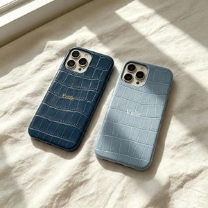 Puede incluir: Dos fundas para smartphone con un patrón de textura de piel de cocodrilo. La funda izquierda es azul marino oscuro con el nombre Frida impreso en dorado. La funda derecha es azul claro con el nombre Viole impreso en blanco. Ambas tienen triple cámara.