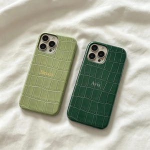 Pode incluir: Duas capas de telemóvel retangulares com um padrão de textura de pele de crocodilo. A capa da esquerda é verde sálvia claro com o nome Becca impresso em dourado. A capa da direita é verde floresta escuro com o nome Avis impresso em prateado.