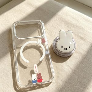 May include: A clear phone case featuring a white magnetic ring, the text MIFFY, and small colorful rabbit illustrations, accompanied by a matching circular phone grip shaped like a white rabbit with black eyes and a cross-shaped mouth.