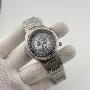 May include: A silver-toned Ricoh world time wristwatch with a stainless steel link bracelet. The dial features a central map of the world, a 24-hour rotating bezel, and a list of global cities. The watch includes a date window at the 3 o'clock position.