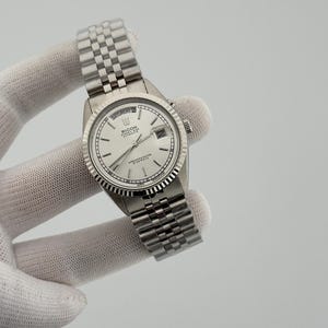 May include: A silver-tone Ricoh automatic wristwatch with a white dial, silver baton indices, and a fluted bezel. The watch features a day-date window at the twelve and three o'clock positions, a stainless steel jubilee-style link bracelet, and a crown.