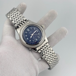Puede incluir: Un reloj de pulsera automático Oris con caja redonda de acero inoxidable, bisel estriado y esfera azul oscuro. El reloj incluye números arábigos blancos, una aguja indicadora de fecha y una pulsera de eslabones de acero inoxidable.