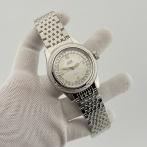 Può includere: Un orologio da polso Oris Automatic color argento con quadrante color crema, numeri arabi a ore 3, 6, 9 e 12, una lancetta puntatore per la data che indica i numeri da 1 a 31 sul bordo esterno e un bracciale a maglie in acciaio inossidabile.