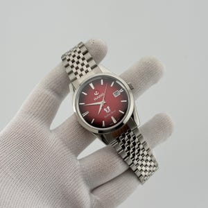 Op de afbeelding: Een Rado Golden Horse polshorloge met een ronde zilverkleurige roestvrijstalen kast en een bijpassende schakelarmband. De wijzerplaat heeft een dieprode gradiëntafwerking, zilverkleurige wijzers, indexen, een datumvenster en een logo.