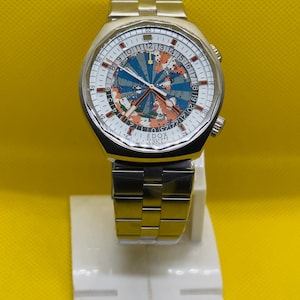 May include: A silver-tone stainless steel wristwatch with a metal link bracelet. The round dial features a colorful world map graphic, a 24-hour scale, and the text Edox Geoscope. The watch has silver hands and two crowns on the right side.