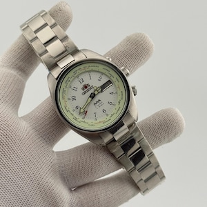 Puede incluir: Un reloj de pulsera Orient de acero inoxidable con esfera blanca, que presenta un bisel de hora mundial con nombres de ciudades, un anillo interior de 24 horas, manecillas luminosas, ventana de día y fecha y una correa de eslabones metálicos.