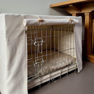 May include: A rectangular metal wire pet crate featuring a cream-coloured fabric cover. The cover includes a roll-up front panel secured with two wooden toggles and loops. Inside the crate, a soft, matching cream-coloured cushion lines the floor.