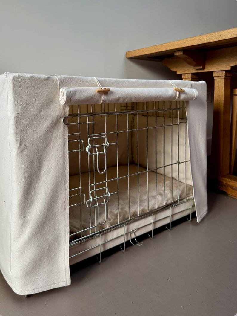May include: A rectangular metal wire dog crate featuring a cream-colored fabric cover. The cover includes a roll-up front panel secured with two wooden toggles and loops. Inside the crate, a soft, matching cream-colored padded mat is visible.