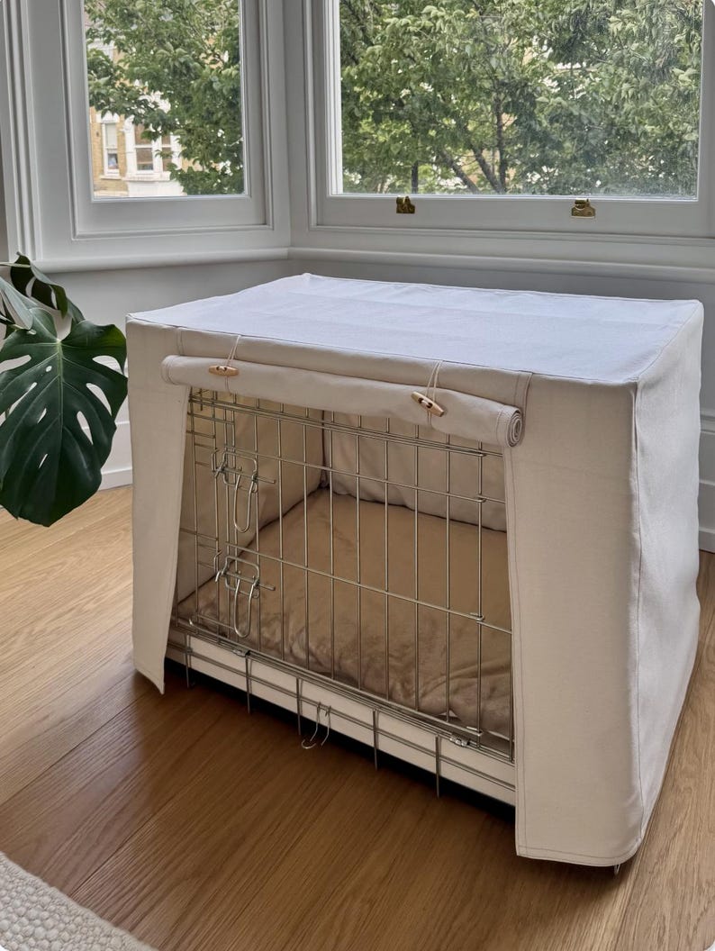 May include: A rectangular metal wire dog crate covered with a solid off-white fabric cover. The front panel of the cover is rolled up and secured with two wooden toggle buttons, revealing the interior metal structure and a tan cushioned floor mat.
