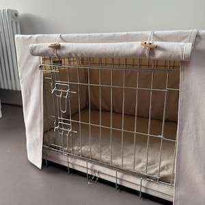May include: A rectangular metal wire dog crate featuring a beige fabric cover. The cover has a roll-up front panel secured by two wooden toggle buttons. Inside the crate, a soft beige mat lines the floor, and the metal frame has two latches.