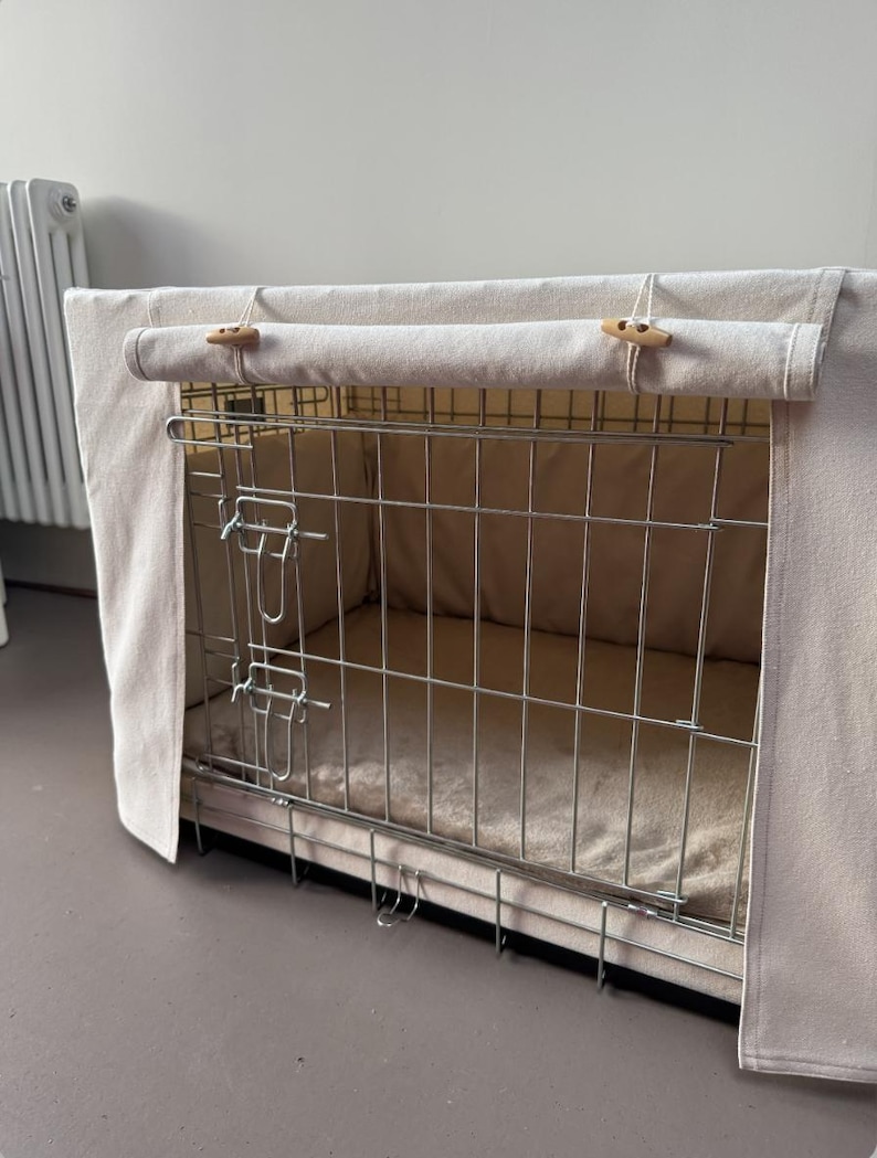 May include: A rectangular metal wire dog crate featuring a beige fabric cover. The cover has a roll-up front panel secured by two wooden toggle buttons. Inside the crate, a soft beige mat lines the floor, and the metal frame has two latches.