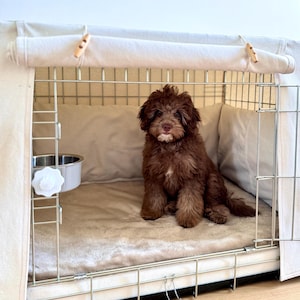 May include: A small, fluffy, dark brown puppy with a white patch on its chin sits inside a wire dog crate. The crate is covered with a beige fabric cover featuring wooden toggles and contains a soft, light-colored plush mat and a metal bowl.