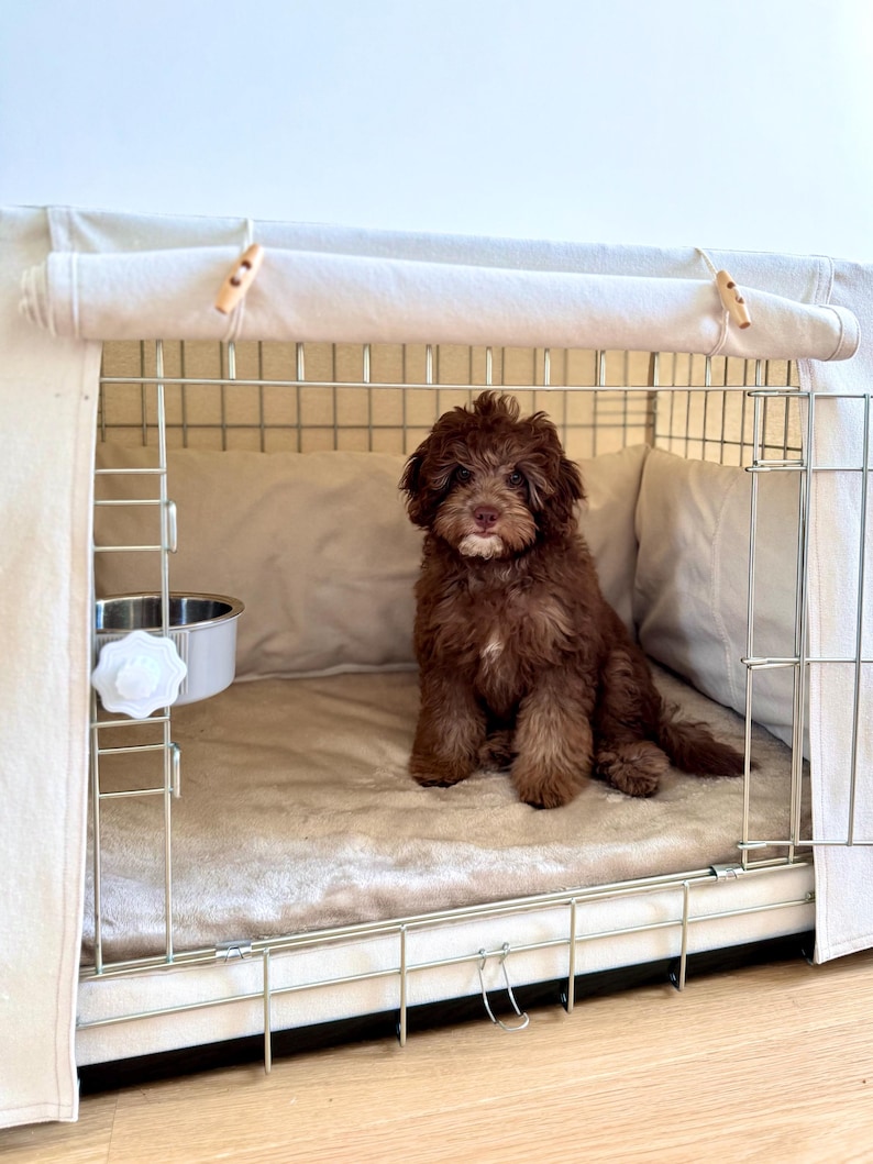 May include: A small, fluffy, dark brown puppy with a white patch on its chin sits inside a wire dog crate. The crate is covered with a beige fabric cover featuring wooden toggles and contains a soft, light-colored plush mat and a metal bowl.