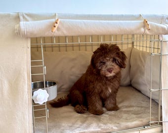 May include: A small, curly-haired brown puppy sits inside a wire dog crate covered with a beige fabric cover. The crate features a soft, plush beige mat on the floor, a stainless steel bowl attached to the side, and wooden toggle fasteners.