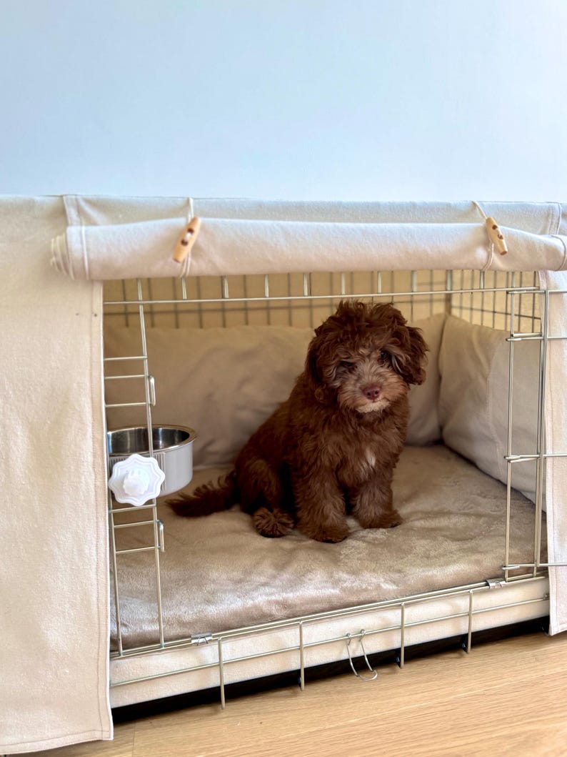 May include: A small, curly-haired brown puppy sits inside a wire dog crate covered with a beige fabric cover. The crate features a soft, plush beige mat on the floor, a stainless steel bowl attached to the side, and wooden toggle fasteners.