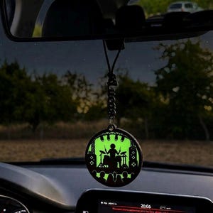 Könnte beinhalten: Ein kreisförmiger Auto-Hängeanhänger mit einer schwarzen Silhouette eines Schlagzeugers, der auf einer Bühne vor einem leuchtend hellgrünen Hintergrund spielt. Der Anhänger hängt an einer schwarzen Kordel und hat ein vertikales Namensschild mit Lucas.