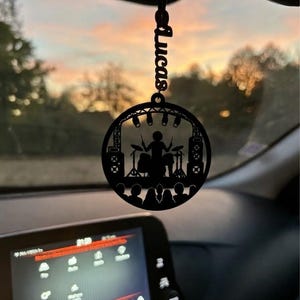 Op de afbeelding: Een zwart rond hangend ornament met een lasergesneden silhouet van een drummer op een podium met podiumverlichting, luidsprekers en een publiek. De naam Lucas is verticaal geïntegreerd in het bovenste bevestigingsstuk.
