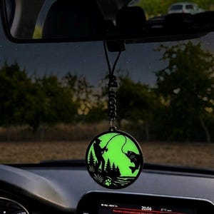 Puede incluir: Un adorno circular colgante para coche con un fondo verde que brilla en la oscuridad y una silueta negra de una persona pescando junto a un bosque con un pez en el anzuelo. El adorno cuelga de un cordón negro con una etiqueta vertical que dice Lucas.