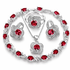 May include: A jewellery set consisting of a bracelet, a ring, a pendant necklace, and two earrings. Each piece features a central round red gemstone surrounded by a textured silver-tone setting encrusted with small clear stones.