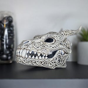 May include: A white and grey dragon skull sculpture featuring intricate, raised knotwork patterns across its surface. The skull has a prominent horn, sharp teeth, and deep eye sockets, with a weathered, textured finish throughout the piece.