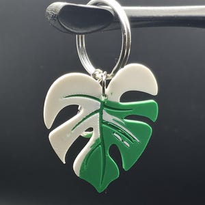 May include: A keychain featuring a split-colored monstera leaf charm. The leaf is divided into two distinct sections, one solid white and one solid green, with characteristic cut-out fenestrations. The charm is attached to a circular silver metal ring.