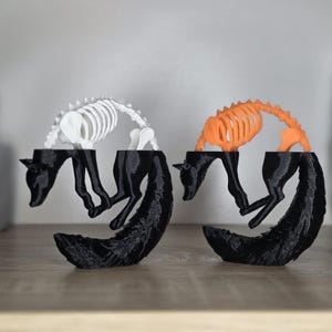 Op de afbeelding: Twee 3D-geprinte vossenbeeldjes in een gekrulde houding. De ene vos heeft een witte skeletachtige ribbenkast en ruggengraat, terwijl de andere een oranje skeletachtige ribbenkast en ruggengraat heeft. Beide vossen hebben zwarte lichamen.