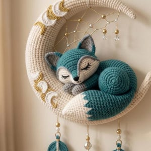 May include: A teal and cream crocheted fox sleeping on a cream-coloured crescent moon. The moon features gold-stitched moon phases, a webbed dreamcatcher design with gold beads, and three hanging teal leaf-shaped ornaments with gold accents.