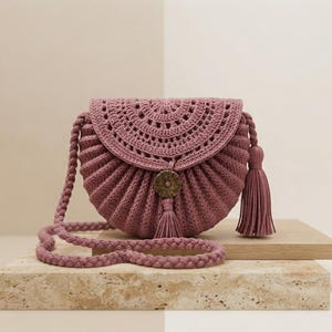 May include: A dusty rose crochet crossbody bag with a semi-circular flap, featuring a textured ribbed body, a braided shoulder strap, and a central wooden button closure. A matching tassel hangs from the bottom of the flap and the side.