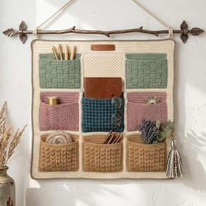Pode incluir: Um organizador de parede em crochê de cor creme, de formato quadrado, com nove bolsos de várias texturas e cores, incluindo verde sálvia, rosa antigo, azul-petróleo escuro e castanho. Está suspenso numa haste decorativa de metal.