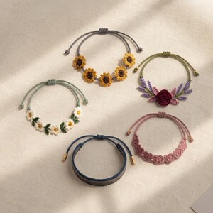 May include: Five adjustable bracelets featuring various designs: one with yellow sunflowers, one with white daisies and green leaves, one with a dark red rose and purple sprigs, one with pink heart-shaped patterns, and one plain dark blue band.