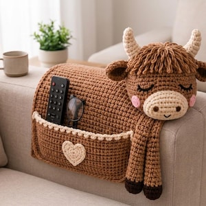 May include: A brown crocheted armrest organizer featuring a highland cow head with cream-colored horns, a fringe of hair, and pink cheeks. The organizer has a front pocket decorated with a cream heart, holding a black remote and eyeglasses.