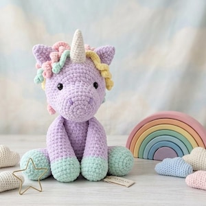 May include: A lavender crochet unicorn plush toy with a white horn, pastel pink, yellow, and mint green yarn mane, and mint green feet. The unicorn is surrounded by small white, blue, and pink crochet clouds, a gold wire star, and a rainbow arch.