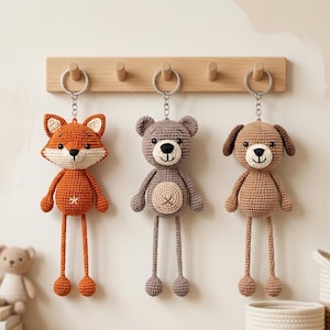 May include: Three crochet keychains hanging from a wooden wall rack. The left keychain is an orange and cream fox, the middle is a grey bear with a cream belly, and the right is a light brown dog. Each features a silver metal ring and chain.