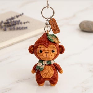 May include: A brown crocheted monkey keychain featuring a small green leaf on its head and a striped green and white scarf around its neck. A brown rectangular tag attached to the metal ring displays the text HANDMADE WITH LOVE in capital letters.