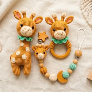 May include: A crochet giraffe toy, a giraffe rattle with a wooden ring, and a pacifier clip featuring a small giraffe head, wooden beads, mint green crocheted spheres, a wooden star, and a wooden moon shape, all crafted in shades of gold and mint.