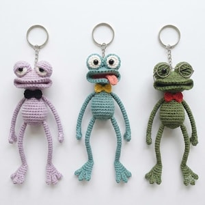 Op de afbeelding: Drie gehaakte kikker-sleutelhangers met metalen ringen. De linker kikker is lichtpaars met een zwarte vlinderdas. De middelste kikker is blauwgroen met een gele vlinderdas en een uitgestoken tong. De rechter kikker is olijfgroen met een rode vlinderdas en een brilletje.