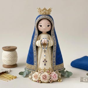 May include: A crocheted doll of a figure wearing a blue and cream gown with a gold crown, holding a rosary. The doll is surrounded by a wooden spool of yarn, a set of gold crochet hooks, a blue fabric square, and a measuring tape showing 7 cm.