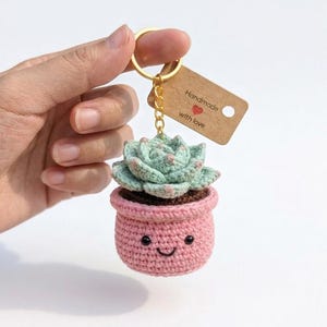 May include: A crochet keychain featuring a light green succulent plant in a pink pot with a stitched smiling face. The keychain has a gold-toned metal ring and a brown rectangular tag with the text Handmade with love and a small red heart.