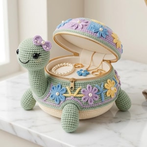 May include: A sage green crocheted turtle jewelry box with a hinged shell lid. The shell is decorated with pastel blue, yellow, and purple crocheted flowers and small white beads. The turtle head features a purple bow and black eyes.