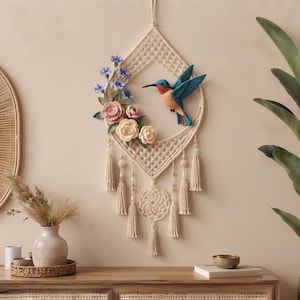 May include: A diamond-shaped macrame wall hanging featuring a blue and orange hummingbird figure, accented with pink, cream, and blue fabric flowers. The piece includes hanging tassels, wooden beads, and a circular woven web detail at the bottom.