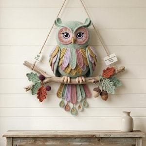 May include: A crocheted owl wall hanging in sage green, dusty pink, and mustard yellow, perched on a natural wooden branch. The owl features large eyes, layered wing feathers, and a long tail. Decorative crocheted oak leaves and acorns hang from the branch, which is suspended by a jute cord. Two small white tags are attached to the cord.